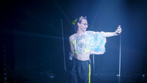 International non-binary beatboxing star SK Shlomo is pictured wearing glasses, a sequin top and black tracksuit trousers. Their arm is outstretched and they look towards their hand which is a holding a phone. 