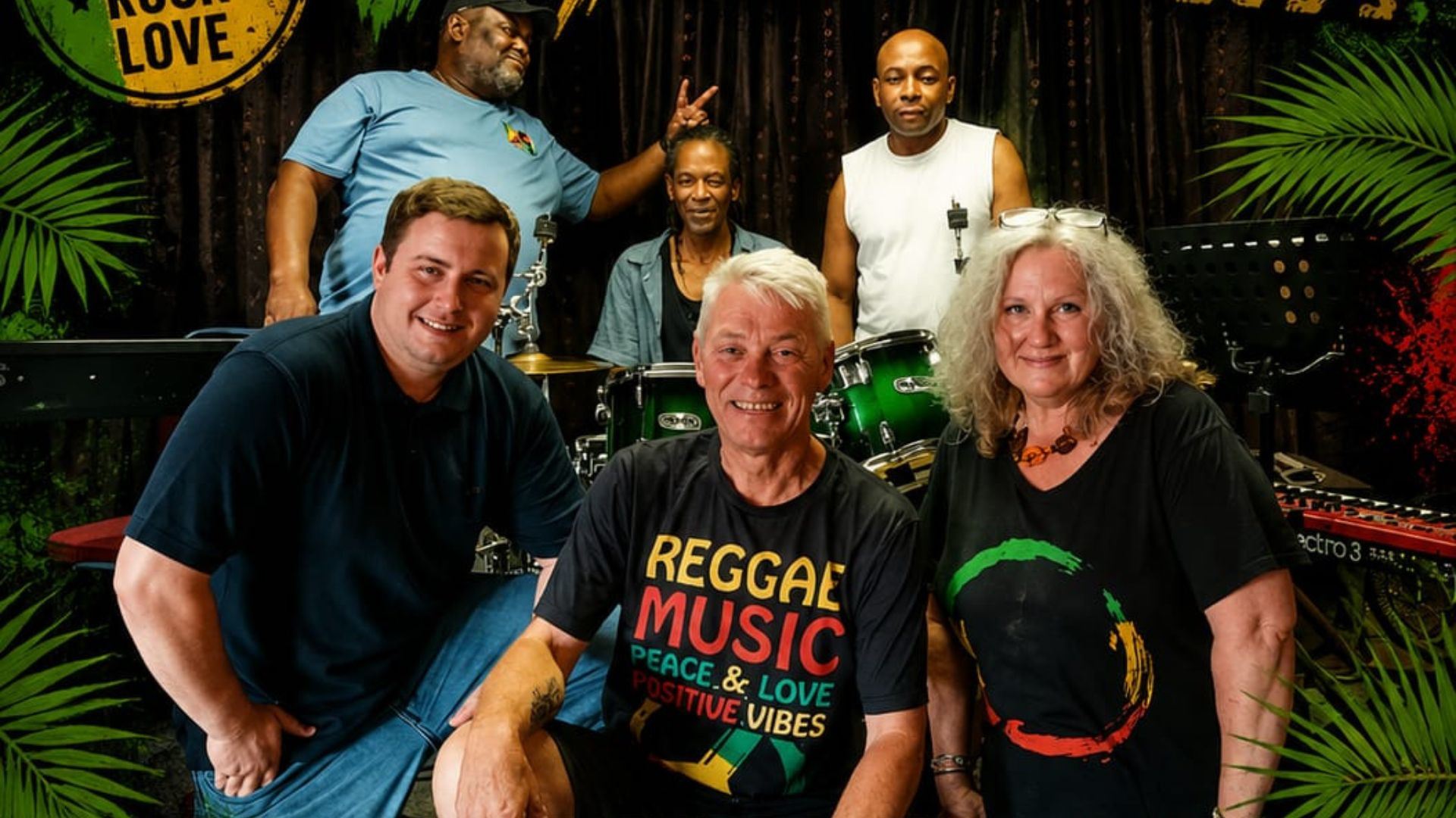 Six people pose together in a music venue with a drum kit and keyboards in the background. Three people stand at the back, while three sit at the front, smiling at the camera. Several wear reggae-themed clothing, and tropical leaf graphics frame the image, creating a vibrant, community-focused atmosphere.
