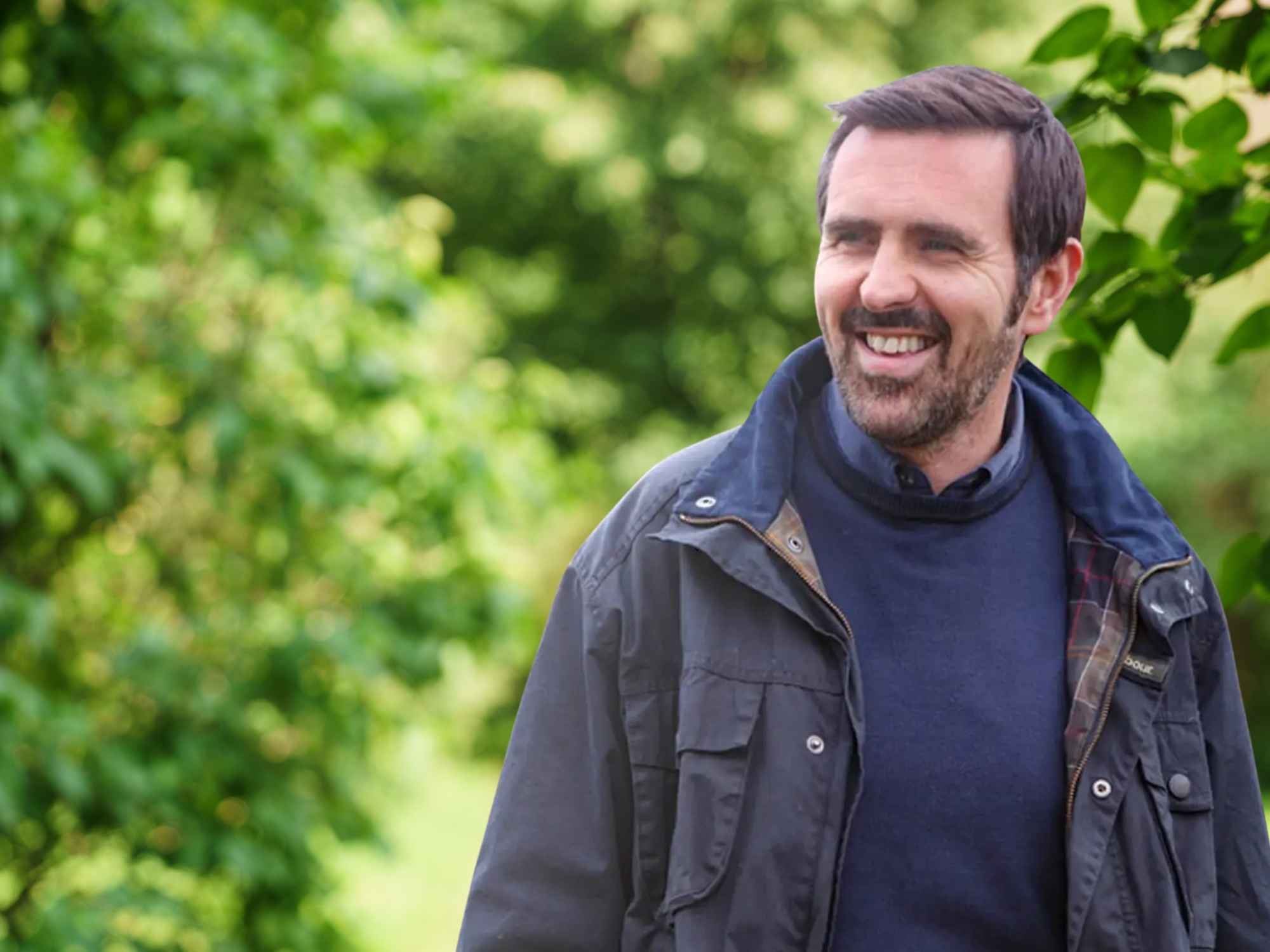 A smiling man with short dark hair and a beard, wearing a navy sweater and outdoor jacket, standing in front of a background of green trees and foliage.