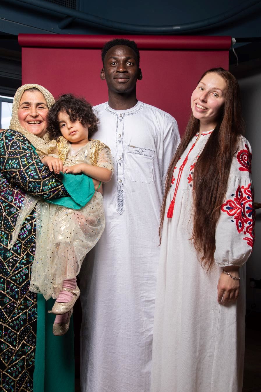 Four people stand together in front of a red backdrop, smiling for a portrait. An older woman wearing a patterned dress and headscarf holds a young child dressed in a light-coloured outfit, while a young man in a white traditional tunic and a woman in a white embroidered dress stand beside them. The group wears a mix of cultural clothing, creating a warm and celebratory atmosphere.